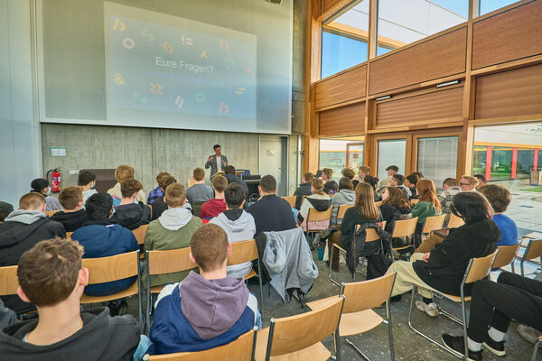 Schüler und Schülerinnen sitzen auf Stühlen in einem Raum, während vorne am Rednerpult jemand einen Vortrag hält.