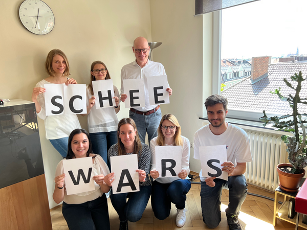 Sieben Mitarbeiterinnen und Mitarbeiter von HeadlineAffairs stehen vor einer Wand und halten jeweils einen Buchstaben auf einem Papier hoch. Als Schriftzug ist "Schee Wars" zu lesen