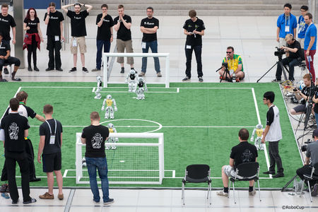kleines Fußballfeld auf dem roboter Fußball spielen. Um das feld herum, stehen viele Menschen und schauen zu.