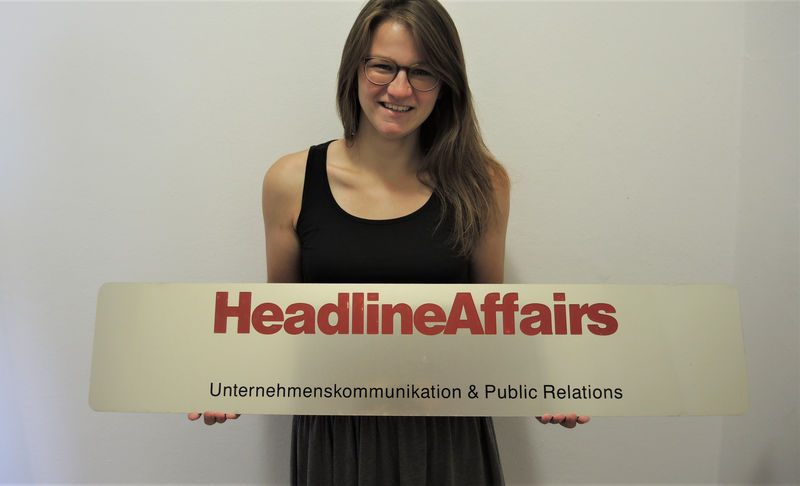 Praktikantin die das HeadlineAffairs Firmenlogo hält. Sie trägt ein schwarzes Top, eine grüne Hose und eine Brille.