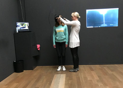 eine Kollegin von HeadlineAffairs, die eine VR Brille von einer Museumsmitarbeiterin aufgezogen bekommt. Beide stehen in einem Raum mit dunkelgrauen Wänden und Holzboden.