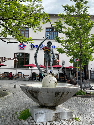 Das Bild zeigt einen kleinen Brunnen, auf dem sich eine Bronzestatue befindet. Die Figur trägt einen Hut aus Bronze und hält verschiedene Blumen in der Hand, die nicht aus Bronze sind. Im Hintergrund sieht man einen Biergarten.