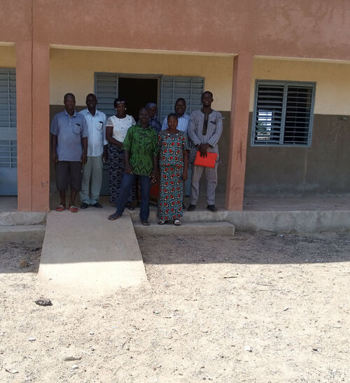 Eine Gruppe von People of Color steht vor einem Gebäude in Burkina Faso und blickt in die Kamera. Vor dem Eingang des Gebäudes liegt Sand. Ein Rollstuhlrampe ist vor der Tür zu sehen.