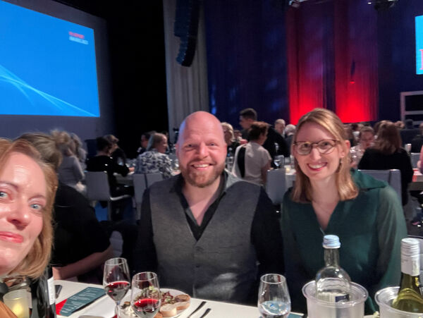 Auf dem Bild sind von links nach rechts Silke Goedereis von Amazon, Christian Bölling von Heldenmood und Ginny Sutter von HeadlineAffairs zu sehen. Sie sitzen an einem Tisch in einem Verleihungssaal und lachen in die Kamera.