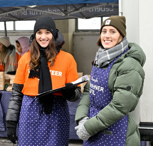 Theresa (rechts) und eine Amazon Mitarbeiterin unterstützen bei der Ausgabe der Spenden bei der Münchner Tafel. Theresa trägt über ihrer Winterjacke eine blaue Schürze mit der Aufschrift "Münchner Tafel" und lacht in die Kamera